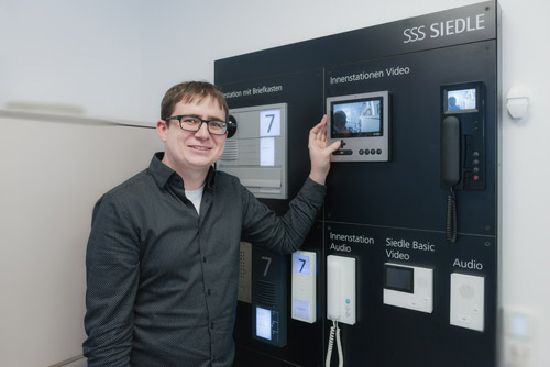 Philipp Denz, ein Mann mit Brille vor einer Wand mit verschiednen Modellen von Sprechanlagen