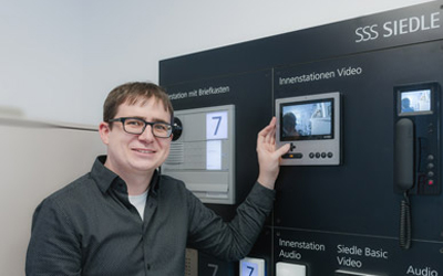 Philipp Denz, ein Mann mit Brille vor einer Wand mit verschiednen Modellen von Sprechanlagen